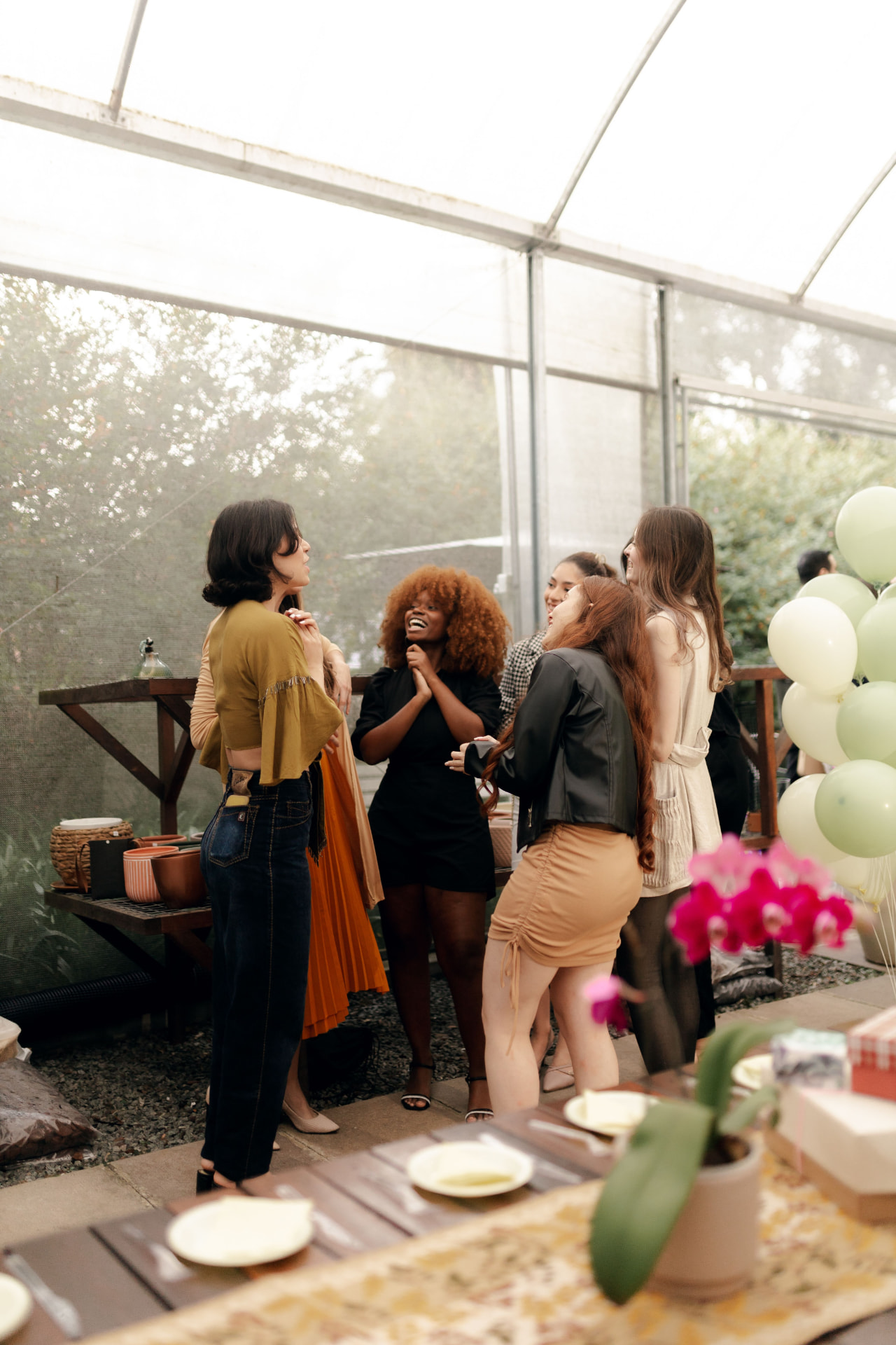 A circle of women laughing together at a gathering in a light-filled greenhouse